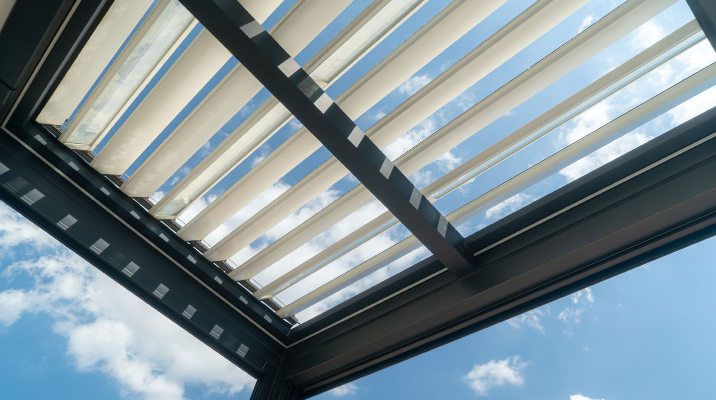 Detalle arquitectónico de lamas de pérgola bioclimática en aluminio gris mostrando tres posiciones de orientación diferentes, con luz natural creando sombras lineales sobre suelo de madera